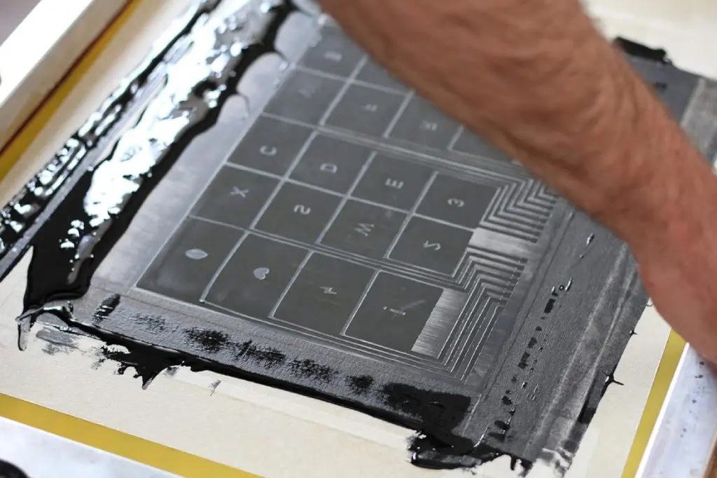 Close-up shot of the screen printing process using black conductive ink to print an intricate electronic circuit with multiple rectangular cells and power symbols on a paper substrate.